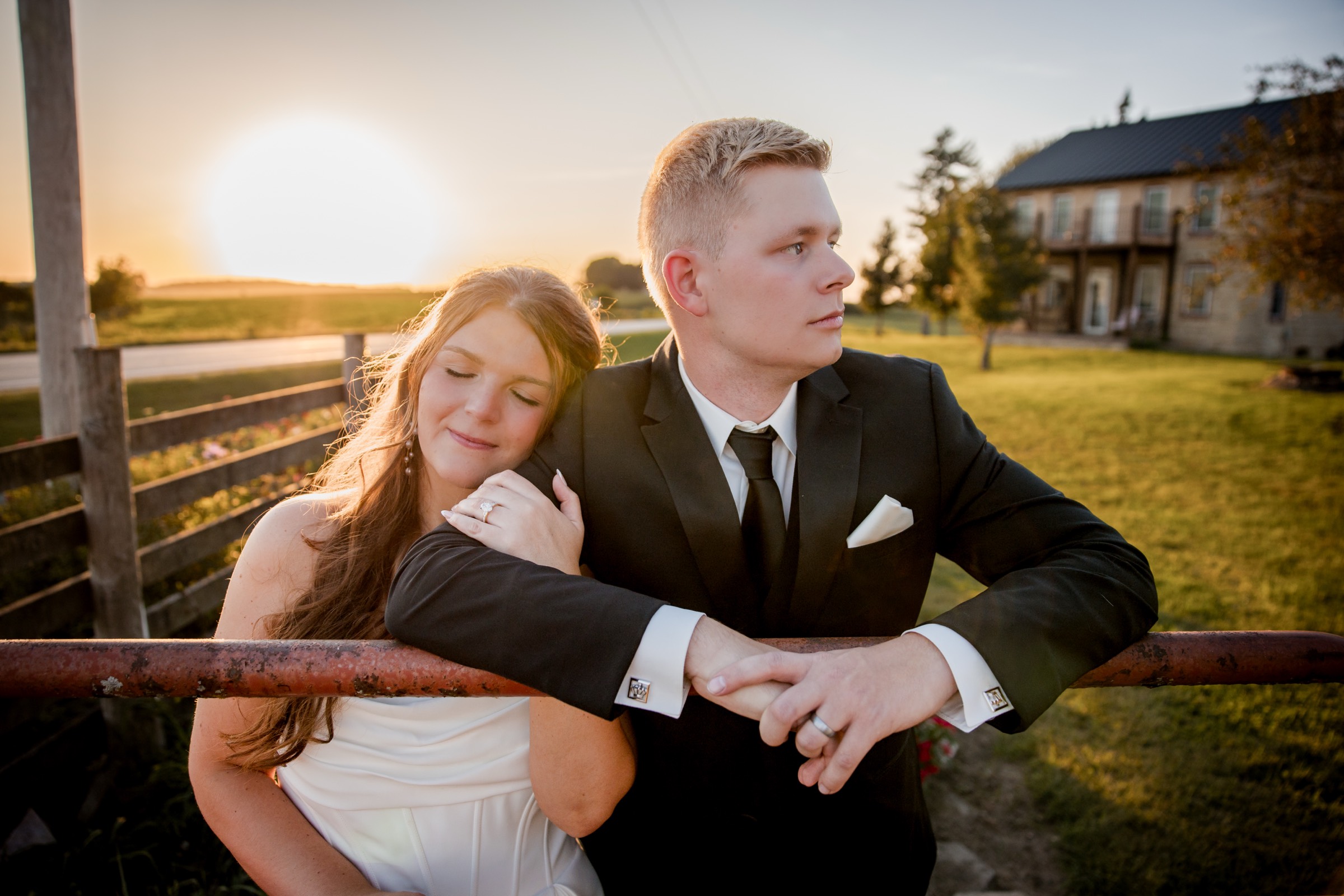 Bride resting her head on the groom's shoulder at a farm gate at sunset
